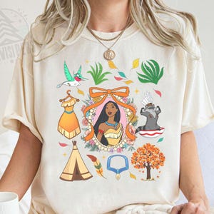 May include: Cream-colored t-shirt featuring a collection of cartoon illustrations. The design includes a portrait of a woman, a hummingbird, a dress, a raccoon, a teepee, and other nature-themed elements. The word "Natural" is printed at the bottom.