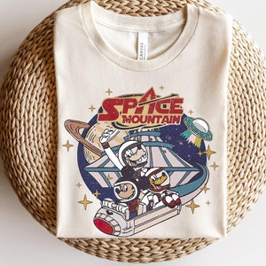 May include: A white t-shirt with a vintage-style graphic of Mickey Mouse, Donald Duck, and Goofy riding a space mountain roller coaster. The graphic includes the text "Space Mountain".