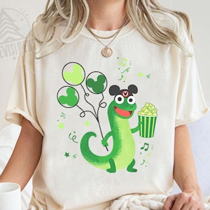 Puede incluir: Camiseta color crema con un dinosaurio verde de dibujos animados sosteniendo globos con forma de Mickey Mouse y un cubo de palomitas de maíz. El dinosaurio lleva un sombrero con orejas de Mickey Mouse. Notas musicales y estrellas decoran el diseño. La palabra "Natural" está impresa en la parte inferior.