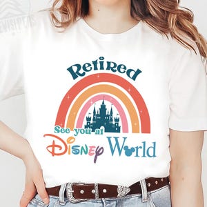K&ouml;nnte beinhalten: Wei&szlig;es T-Shirt mit einem Regenbogen-Design und dem Text "Retired See you at Disney World".