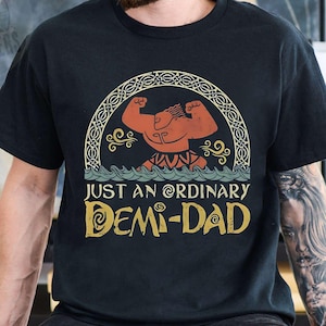 Op de afbeelding: Zwart T-shirt met een vintage-stijl illustratie van een gespierd figuur met een kroon en de tekst "Just an Ordinary Demi-Dad".