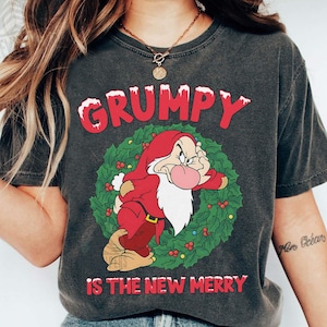 Puede incluir: Camiseta gris oscuro con un gráfico de Gruñón de Blancanieves, rodeado por una corona, y el texto "GRUMPY IS THE NEW MERRY" en rojo y blanco. Un estilo casual y cómodo.