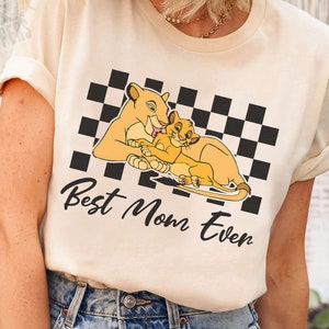 May include: A light beige t-shirt with a black and white checkered pattern. The shirt features a cartoon image of a lioness and a lion cub with the text "Best Mom Even".
