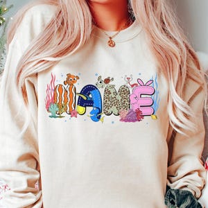 May include: Beige sweatshirt with the word "NAME" in colorful, cartoon-style lettering. The letters are designed with ocean-themed elements, including fish, coral, and a starfish. The sweatshirt has long sleeves.
