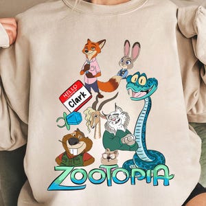 Puede incluir: Sudadera beige de cuello redondo con un gr&aacute;fico colorido de personajes de la pel&iacute;cula animada "Zootopia". El dise&ntilde;o incluye un zorro, un conejo, una serpiente y otros animales, con la palabra "Zootopia" en verde y azul. Una etiqueta con el nombre dice "Hello Clark."