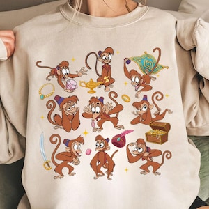 May include: Beige sweatshirt featuring a cartoon design of a monkey character in various poses. The design includes a magic lamp, treasure chest, and a magic carpet. The monkey is depicted in shades of brown, with accents of gold, blue, and red.