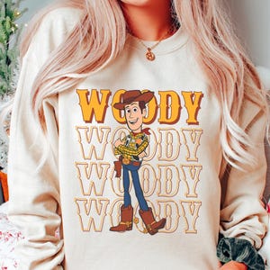 Puede incluir: Una sudadera beige con un gr&aacute;fico del personaje Woody de Toy Story. La palabra "Woody" est&aacute; impresa en letras grandes de color naranja encima del personaje, con el nombre repetido debajo. Woody est&aacute; representado con su atuendo de vaquero caracter&iacute;stico.