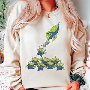 May include: A beige sweatshirt featuring a cartoon design of the Little Green Men from Toy Story being lifted by a rocket claw. The aliens are green with three eyes and blue uniforms. The rocket is blue and green.