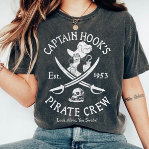 May include: A dark gray t-shirt with a white graphic of Captain Hook's pirate crew. The graphic includes a skull and crossbones, the text "Captain Hook's", "Est. 1953", "Pirate Crew", and the phrase "Look Alive, You Swabs!"