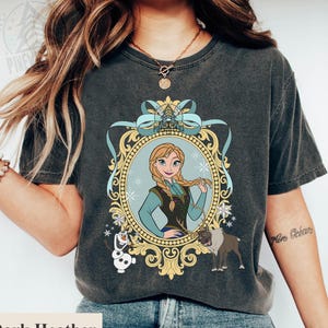 Könnte beinhalten: Dunkelgraues Heather-T-Shirt mit einem gerahmten Cartoon-Porträt einer Figur mit blonden Zöpfen, die ein teal-schwarzes Outfit trägt. Der goldene Rahmen hat eine blaue Schleife. Enthält eine Schneemann- und Rentier-Grafik.