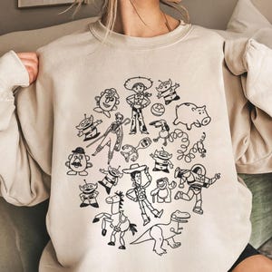 Può includere: Felpa girocollo beige con un disegno a tratto nero di vari personaggi dei cartoni animati. Il design include un cowboy, alieni, un maiale e altre figure. La felpa è realizzata in un materiale morbido.