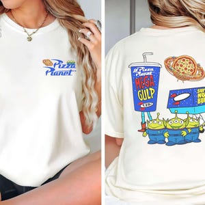 May include: Cream-colored t-shirt featuring the Pizza Planet logo on the front. The back displays a Mega Gulp cup, pizza, a Super Nova Burger box, and three green aliens in blue jumpsuits. A fun, cartoon-style design.