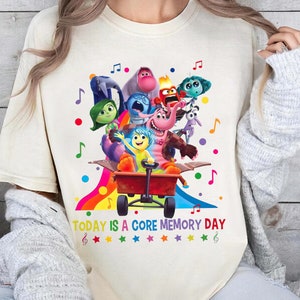 May include: A white t-shirt featuring a colourful graphic of characters from the film Inside Out. The characters are riding in a red wagon. The text "TODAY IS A CORE MEMORY DAY" is printed below the graphic.