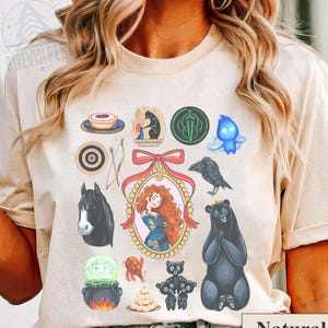 May include: Beige t-shirt featuring a collection of colorful illustrations from a movie. The design includes a portrait, a black bear, a raven, and other themed graphics. The word "Natural" is printed at the bottom.
