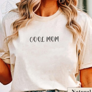 May include: A cream-colored t-shirt with the words "COOL MOM" printed in black script. The shirt has short sleeves and a crew neck. The word "Natural" is printed in a white rectangle at the bottom.