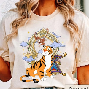 May include: A beige t-shirt featuring a colorful graphic of a tiger, monkey, magic lamp, and magic carpet. The tiger is orange with black stripes. The monkey is brown. The shirt has the word "Natural" printed on the bottom.