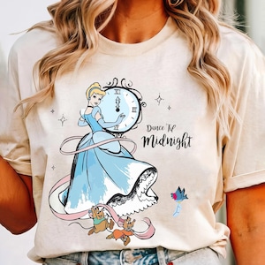 Può includere: Una maglietta beige chiaro con una grafica di Cenerentola in un abito blu che tiene un orologio con il testo "Dance 'Til Midnight".