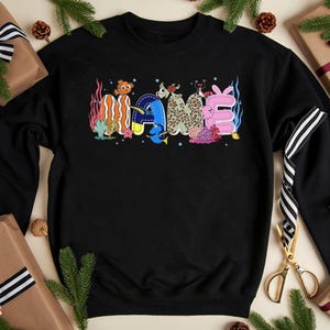 May include: Black sweatshirt with the word "NAME" in colorful, whimsical lettering. The letters are decorated with sea creatures and coral, creating a playful, underwater theme. A fun, casual top.