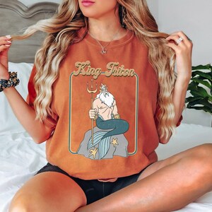 May include: A brown t-shirt featuring a retro-style graphic of King Triton from The Little Mermaid. The graphic depicts King Triton sitting on a rock with his trident and a blue and green tail.