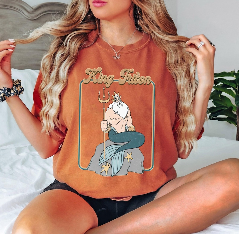 May include: A brown t-shirt featuring a retro-style graphic of King Triton from The Little Mermaid. The graphic depicts King Triton sitting on a rock with his trident and a blue and green tail.