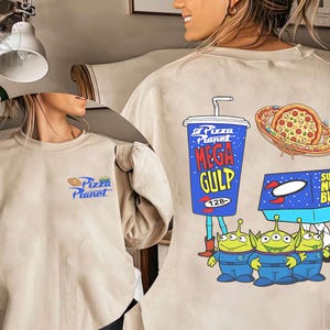 May include: Beige sweatshirt featuring a Pizza Planet logo on the front. The back displays a Mega Gulp cup, pizza, Super Nova Burger box, and three green alien characters. The design is in a cartoon style.