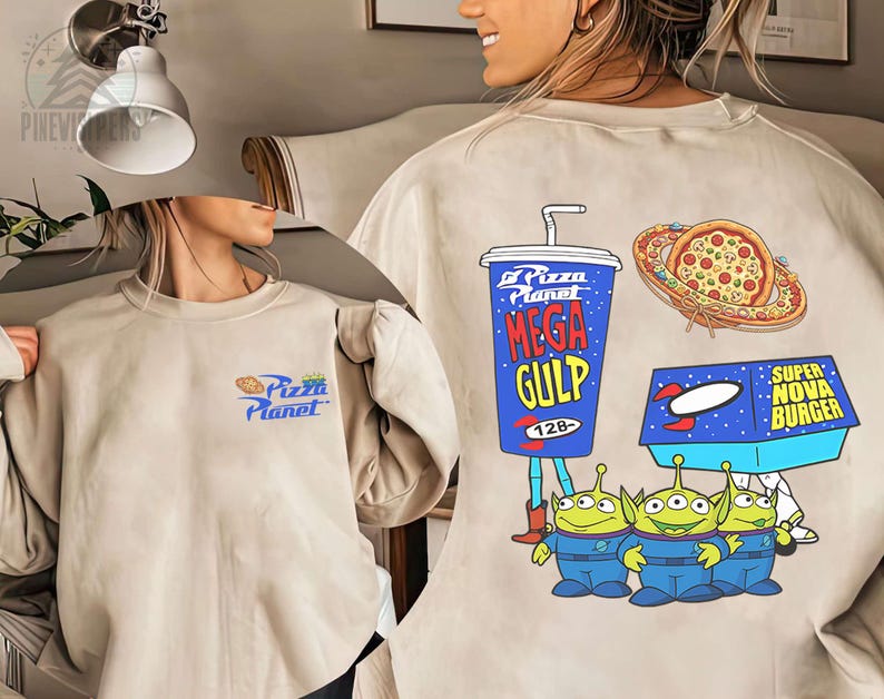 May include: Beige sweatshirt featuring a Pizza Planet logo on the front. The back displays a Mega Gulp cup, pizza, Super Nova Burger box, and three green alien characters. The design is in a cartoon style.