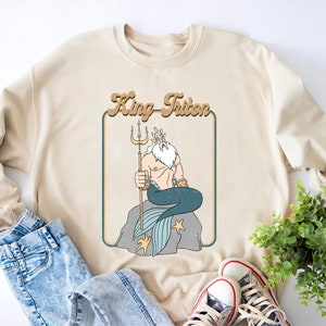 May include: A beige sweatshirt featuring a retro-style graphic of King Triton from The Little Mermaid. The graphic depicts King Triton sitting on a rock with his trident and a blue tail.
