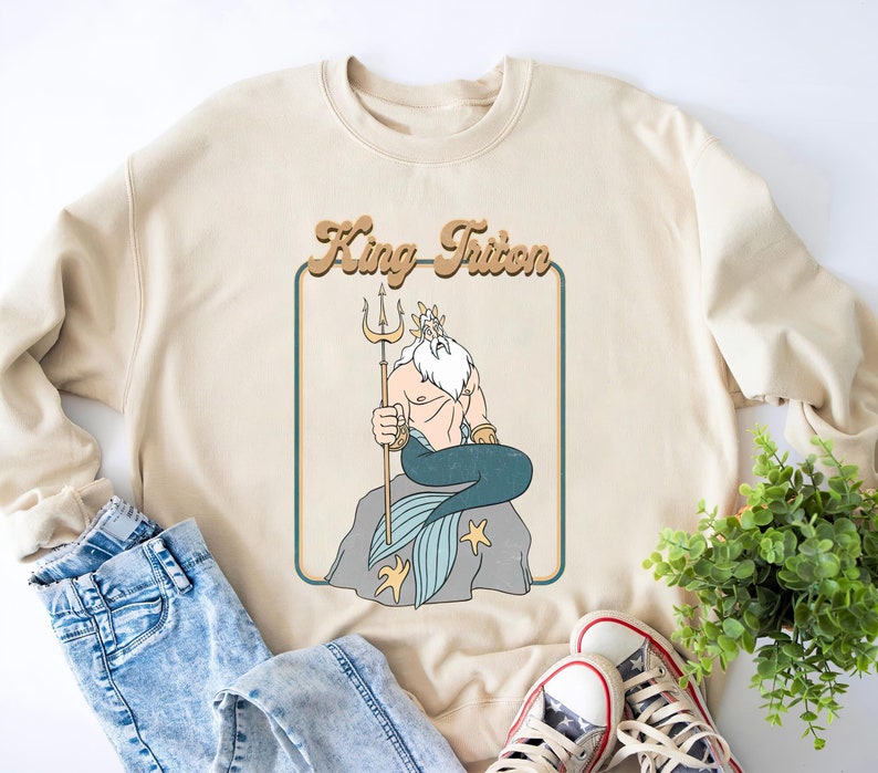 May include: A beige sweatshirt featuring a retro-style graphic of King Triton from The Little Mermaid. The graphic depicts King Triton sitting on a rock with his trident and a blue tail.