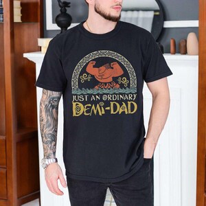 Op de afbeelding: Zwart T-shirt met een vintage-stijl grafische afbeelding van een gespierd cartoonfiguur met de tekst "Just an ordinary Demi-Dad".