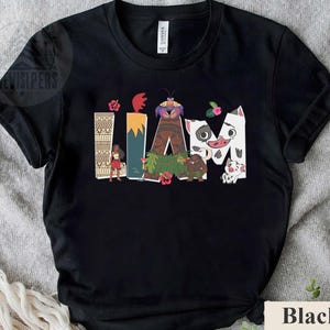May include: Black t-shirt with the word "IAM" in colourful, stylised letters, featuring cartoon characters and tropical floral accents. The design includes a woman, a crab, and a pig. The shirt is made of a soft material.