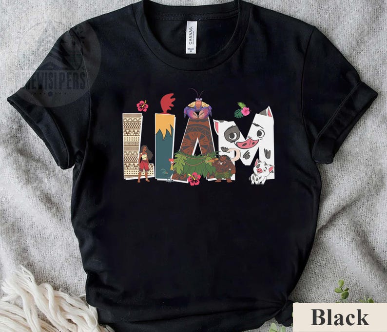 May include: Black t-shirt with the word "IAM" in colourful, stylised letters, featuring cartoon characters and tropical floral accents. The design includes a woman, a crab, and a pig. The shirt is made of a soft material.