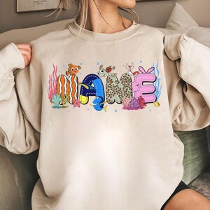 May include: Beige sweatshirt with the word "MAME" in colorful, cartoonish letters. The letters are designed with aquatic themes, including fish, coral, and bubbles. The sweatshirt has long sleeves and a crew neck.