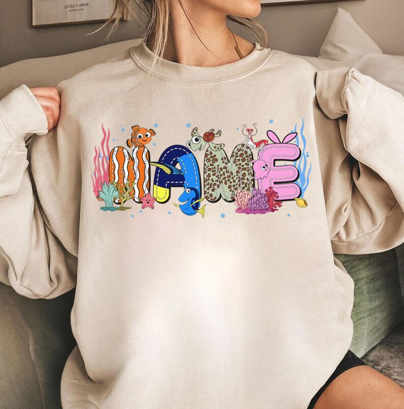May include: Beige sweatshirt with the word "MAME" in colorful, cartoonish letters. The letters are designed with aquatic themes, including fish, coral, and bubbles. The sweatshirt has long sleeves and a crew neck.