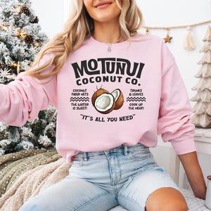 May include: A light pink sweatshirt with the text "MOTUNUI COCONUT CO." and a graphic of a coconut. The sweatshirt also features text about coconut fiber nets, trunks & leaves, and the phrase "IT'S ALL YOU NEED".