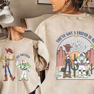 May include: Beige sweatshirt featuring cartoon characters Woody and Buzz Lightyear from Toy Story. The front displays Woody and Buzz with the text "Big Brother." The back has a desert scene with the text "You've Got a Friend in Me."