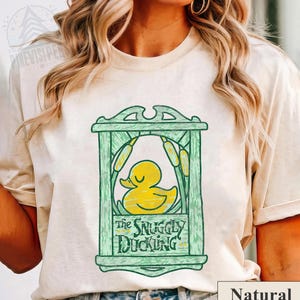 May include: A natural-colored t-shirt featuring a green frame design with a yellow duck illustration. The text "The Snuggly Duckling" is below the duck. The shirt has a relaxed fit and short sleeves.