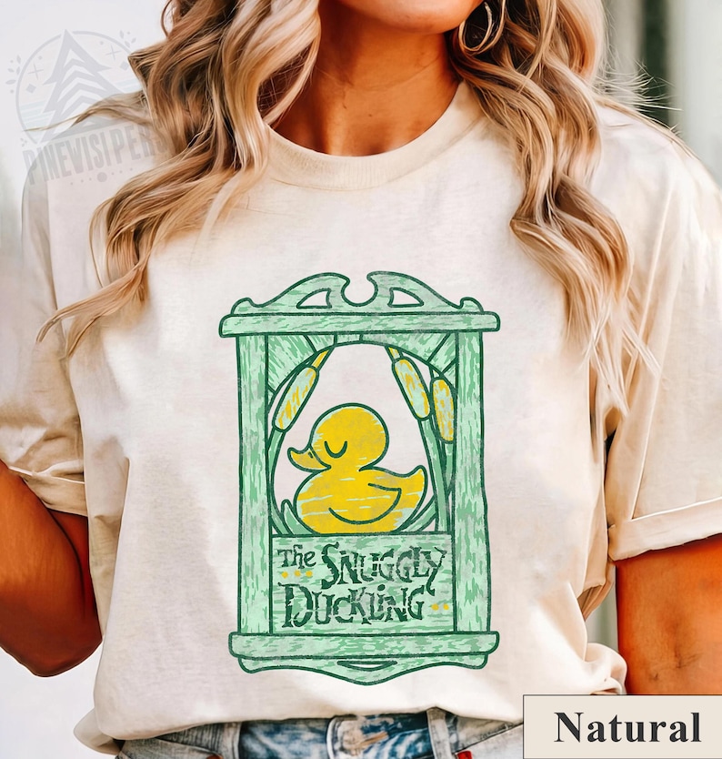 May include: A natural-colored t-shirt featuring a green frame design with a yellow duck illustration. The text "The Snuggly Duckling" is below the duck. The shirt has a relaxed fit and short sleeves.
