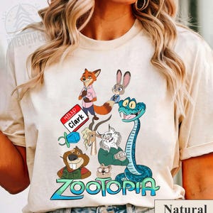 Puede incluir: Camiseta de color natural con un gr&aacute;fico colorido de personajes de la pel&iacute;cula animada "Zootopia". El dise&ntilde;o incluye un zorro, un conejo, una serpiente y otros personajes de animales, junto con el t&iacute;tulo de la pel&iacute;cula en azul y verde.
