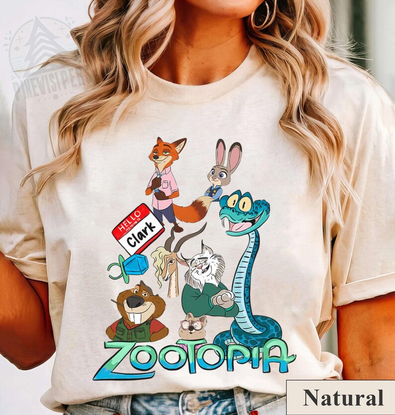 Puede incluir: Camiseta de color natural con un gr&aacute;fico colorido de personajes de la pel&iacute;cula animada "Zootopia". El dise&ntilde;o incluye un zorro, un conejo, una serpiente y otros personajes de animales, junto con el t&iacute;tulo de la pel&iacute;cula en azul y verde.