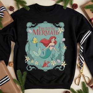 May include: Black sweatshirt featuring a graphic of Ariel from "The Little Mermaid." The design includes Ariel with red hair and a teal tail, along with Flounder and Sebastian. The words "The Little Mermaid" are at the top.