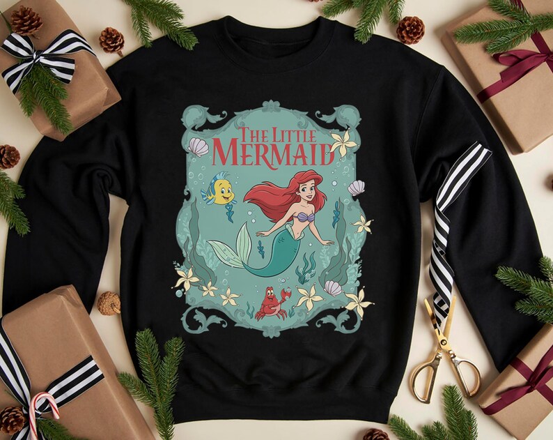 May include: Black sweatshirt featuring a graphic of Ariel from "The Little Mermaid." The design includes Ariel with red hair and a teal tail, along with Flounder and Sebastian. The words "The Little Mermaid" are at the top.