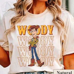 Puede incluir: Camiseta color crema con un gr&aacute;fico de Woody de Toy Story. El dise&ntilde;o incluye la palabra "WOODY" en naranja, repetida verticalmente, y una ilustraci&oacute;n del personaje con su atuendo de vaquero. La palabra "Natural" est&aacute; en un cuadro blanco en la parte inferior.