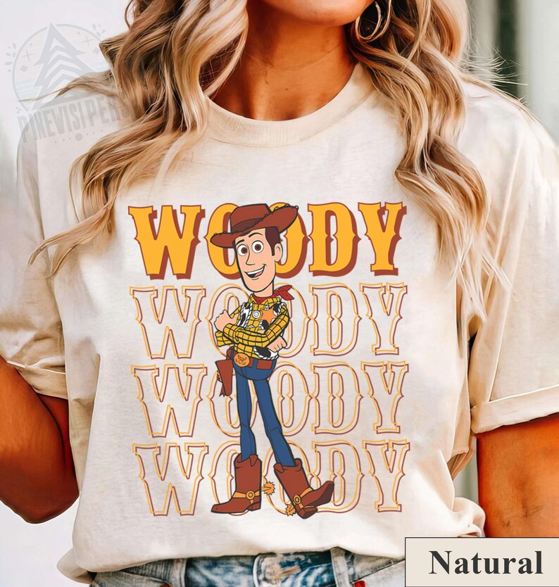 Puede incluir: Camiseta color crema con un gr&aacute;fico de Woody de Toy Story. El dise&ntilde;o incluye la palabra "WOODY" en naranja, repetida verticalmente, y una ilustraci&oacute;n del personaje con su atuendo de vaquero. La palabra "Natural" est&aacute; en un cuadro blanco en la parte inferior.