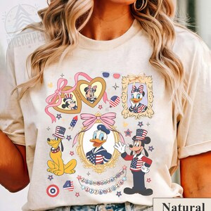 Könnte beinhalten: Beiges T-Shirt mit patriotischen Disney-Figuren, darunter Mickey, Minnie, Donald und Goofy, in Rot, Weiß und Blau. Das Design umfasst gerahmte Porträts und den Text "250th Anniversary 1776-2026".