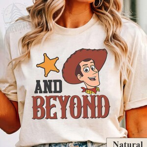 May include: A natural-colored t-shirt featuring a cartoon image of a cowboy with a sheriff's star and the words "AND BEYOND" in a western-style font. The shirt has a crew neck and short sleeves.