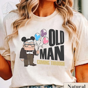 Puede incluir: Camiseta beige con un dibujo animado de un anciano con orejas de Mickey Mouse sosteniendo globos. El texto "OLD MAN COMING THROUGH" está impreso en negro y dorado. La camiseta está etiquetada como "Natural".