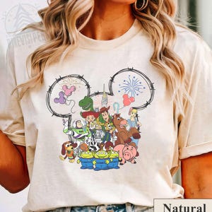 Pu&ograve; includere: T-shirt beige con una grafica colorata dei personaggi di Toy Story della Disney all'interno di un contorno della testa di Topolino. Il design include personaggi come Woody, Buzz Lightyear e i Little Green Men. La parola "Natural" &egrave; stampata in basso.