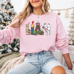 May include: A pale pink sweatshirt featuring the name "LIAM" in a colourful, cartoon design. The letters are decorated with images of characters and flowers. The sweatshirt has long sleeves and is a casual garment.