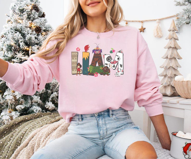 May include: A pale pink sweatshirt featuring the name "LIAM" in a colourful, cartoon design. The letters are decorated with images of characters and flowers. The sweatshirt has long sleeves and is a casual garment.