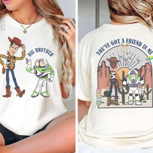 May include: Cream-colored t-shirt featuring cartoon characters Woody and Buzz Lightyear. The front of the shirt has the text "BIG BROTHER". The back of the shirt has the text "YOU'VE GOT A FRIEND IN ME".